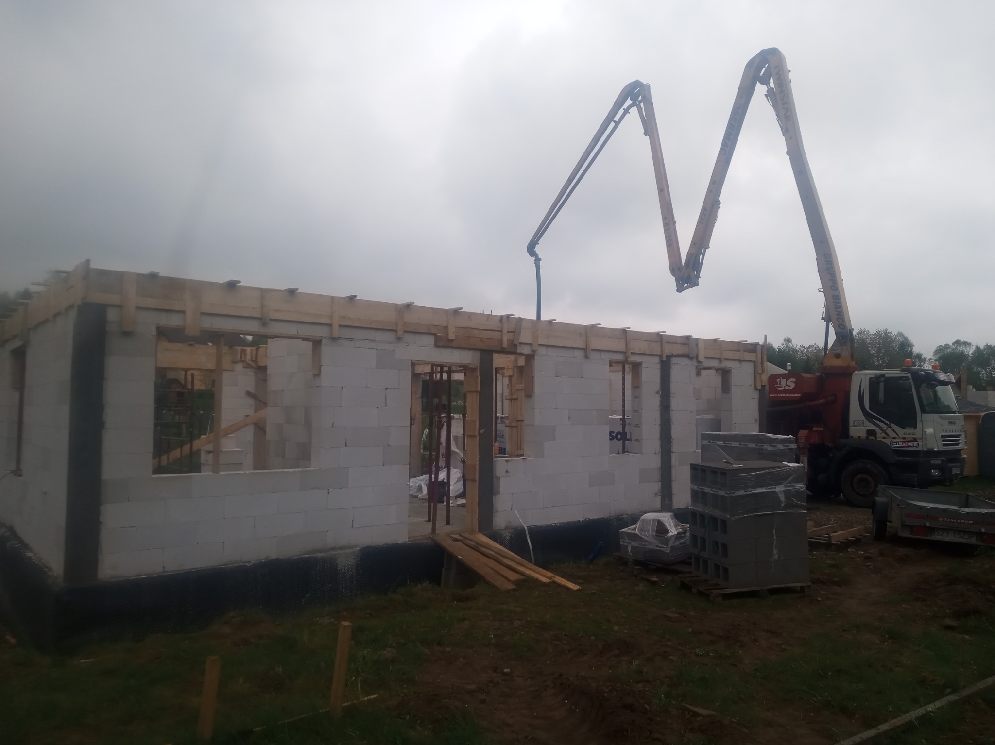 Wznoszenie ścian parterowego domu z bloczków betonowych, widoczna pompa do betonu i drewniane szalunki pod wieniec, pochmurne niebo w tle.