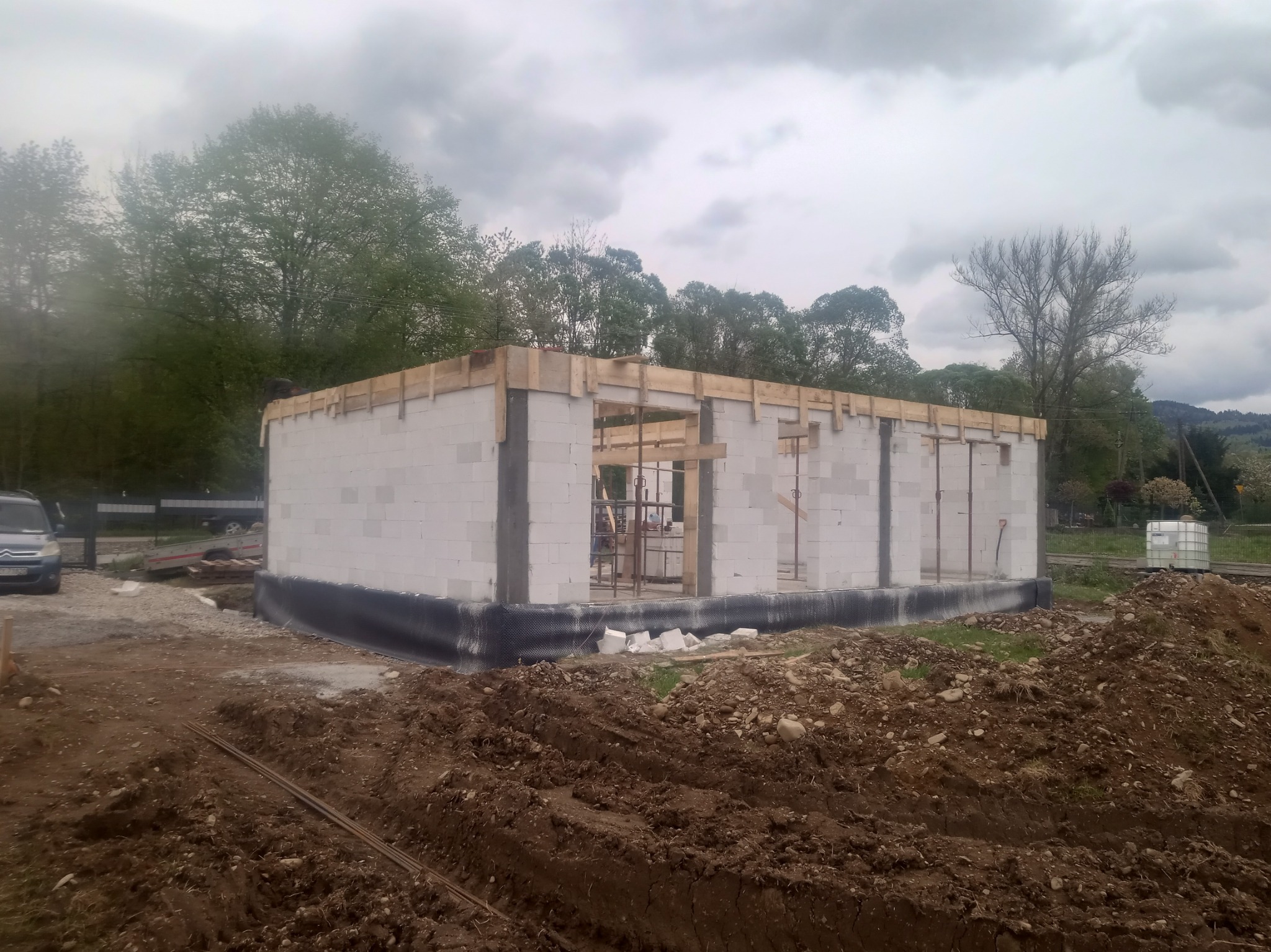 Budowa domu z białych bloczków, widoczne szalunki na strop, teren wokół w stanie surowym, pochmurne niebo w tle.