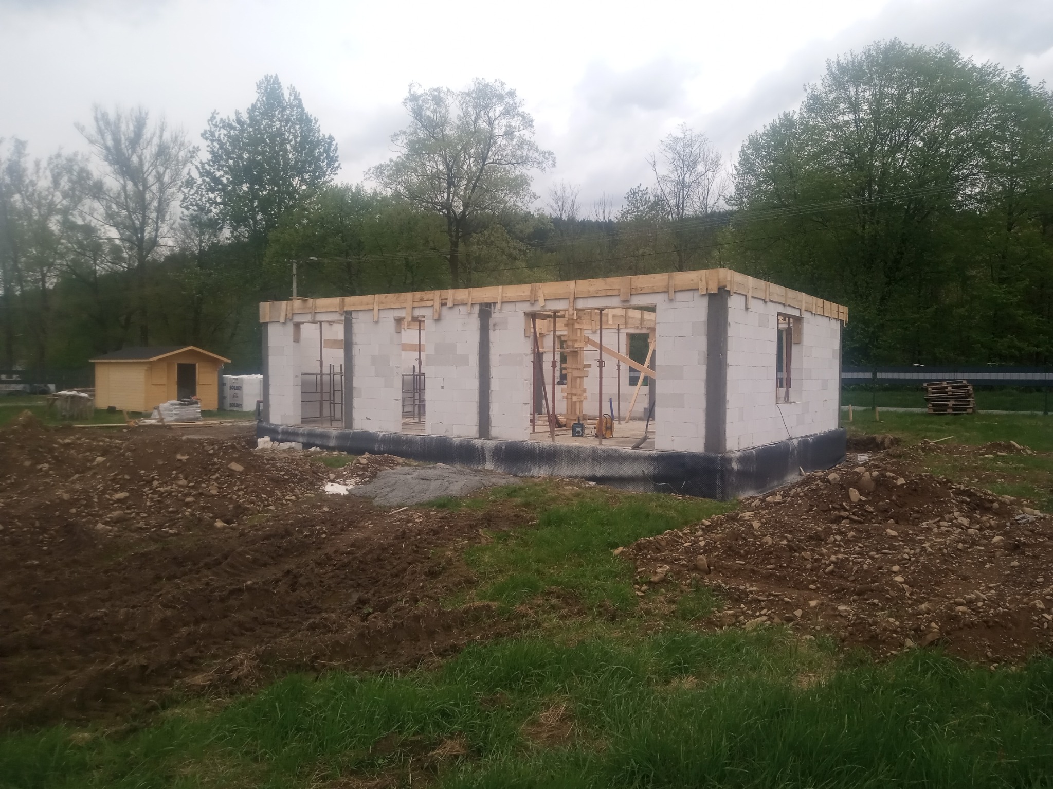 Stan surowy otwarty domu murowanego w Węgierskiej Górce z widocznym szalunkiem na strop i przygotowanym terenem wokół, w tle mała drewniana altanka.