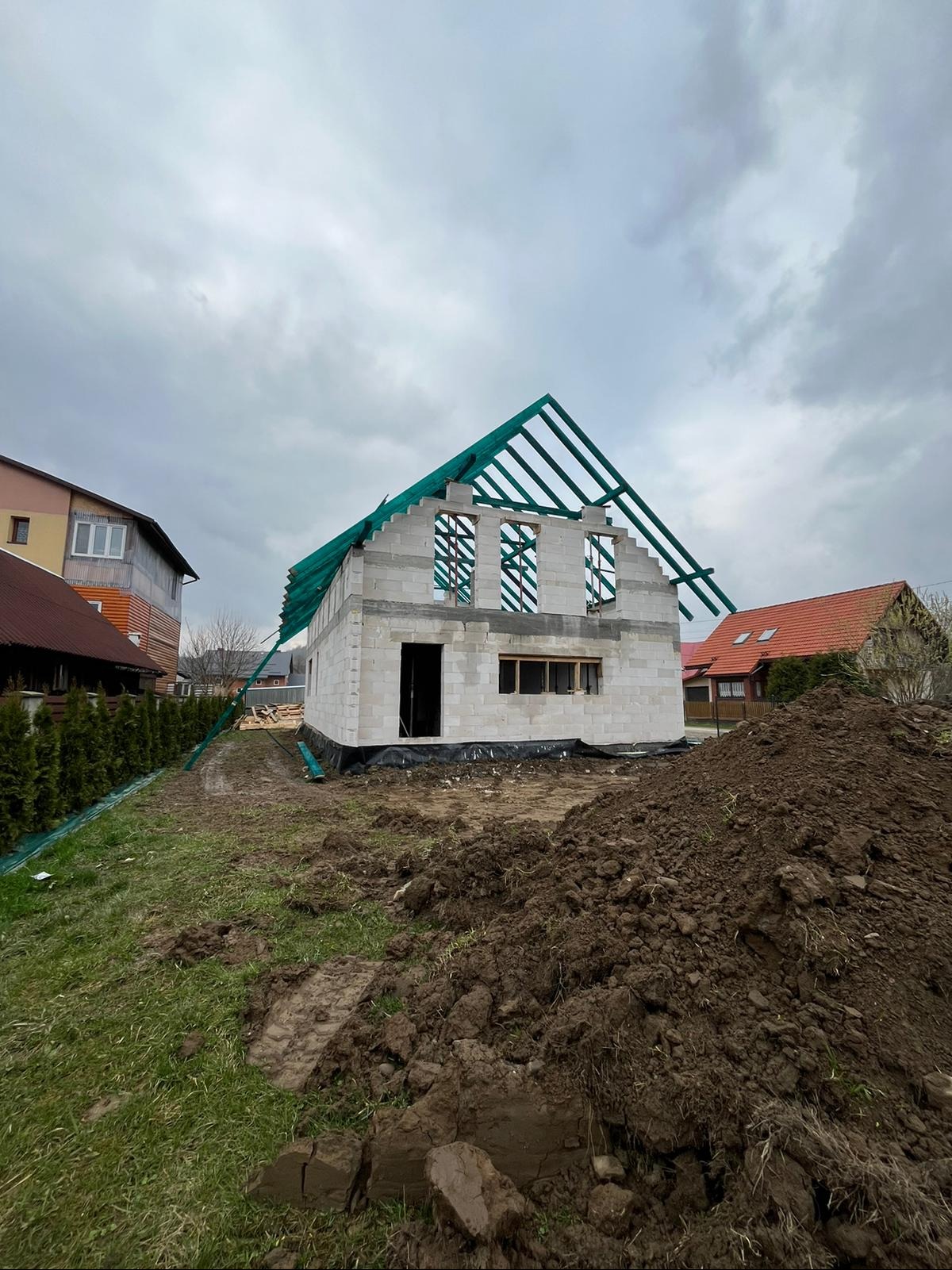 Surowy stan budowy domu z białych bloczków, widoczna konstrukcja dachu z zieloną folią, rozkopana ziemia w pobliżu, pochmurne niebo w tle.