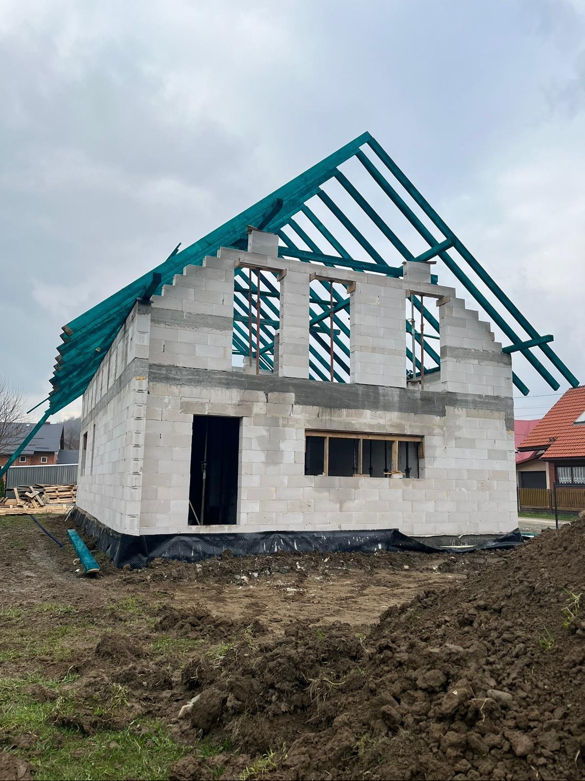 Surowy stan domu z białych bloczków, widoczne okna, drzwi i zielona konstrukcja dachu na tle pochmurnego nieba, ziemia przed budynkiem częściowo rozkopana.