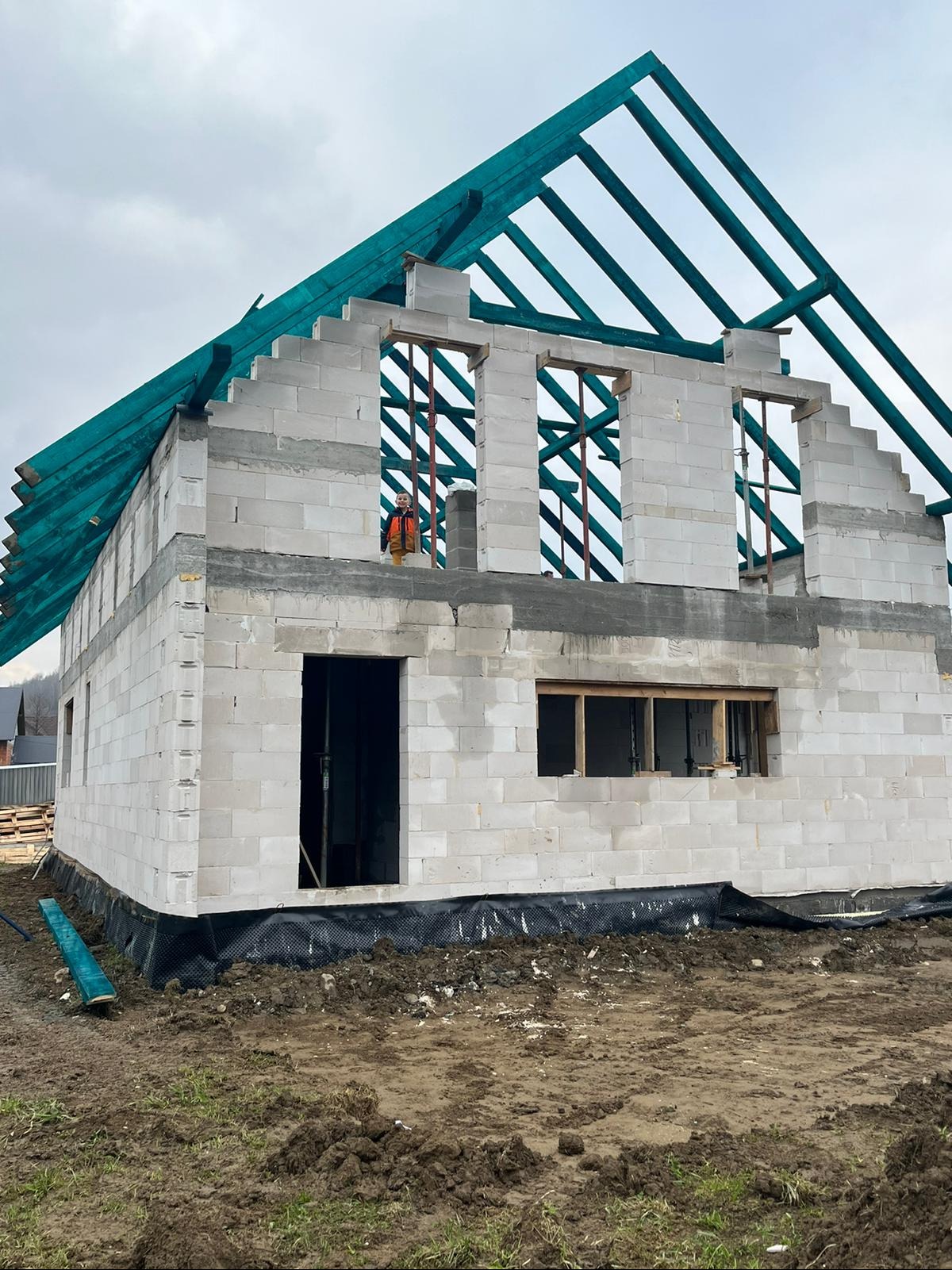 Surowy stan domu z betonu komórkowego w trakcie budowy. Widoczne ściany, otwory okienne z drewnianymi ramami, zielona konstrukcja dachu i pracownik w pomarańczowej kamizelce na górze.