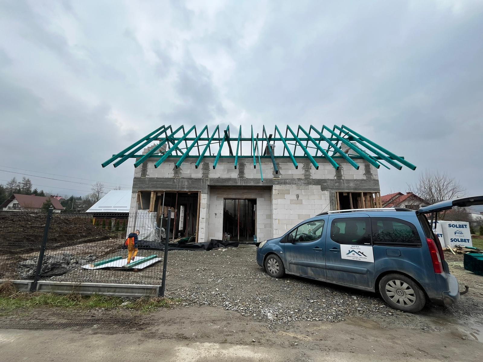 Wznoszony dom murowany z widoczną konstrukcją dachu z zielonych belek, zaparkowany obok niebieski samochód dostawczy z logo firmy budowlanej.