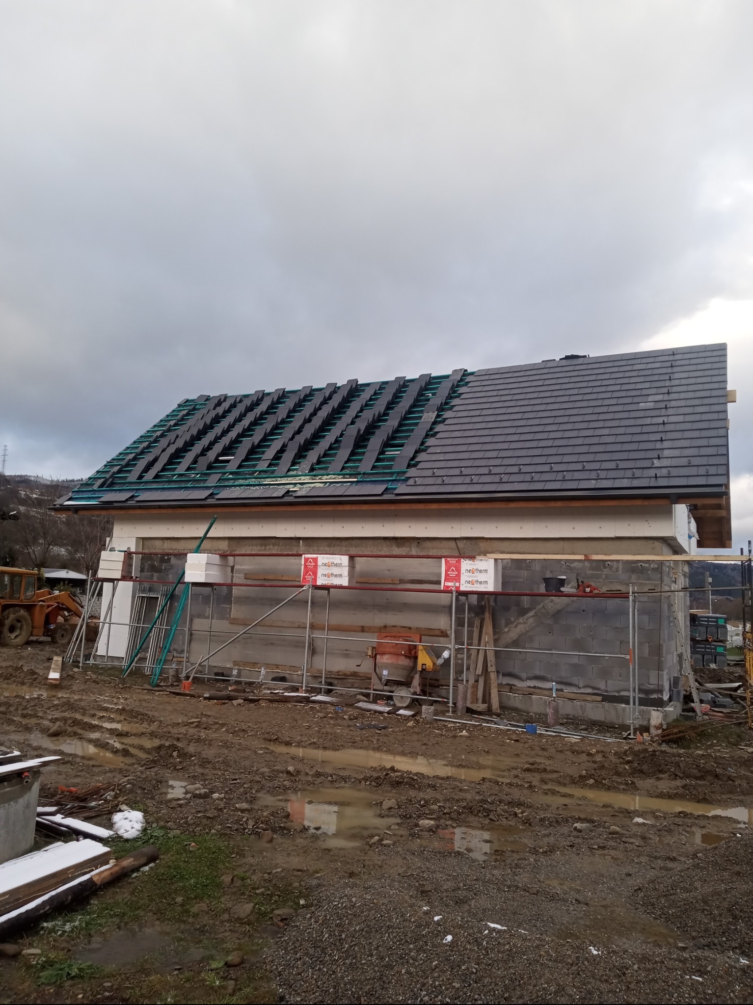 Budowa domu murowanego w trakcie układania dachówki; część dachu pokryta, na drugiej części widoczne membrany i łaty; na pierwszym planie rusztowania i materiały budowlane, teren wokół placu budowy...