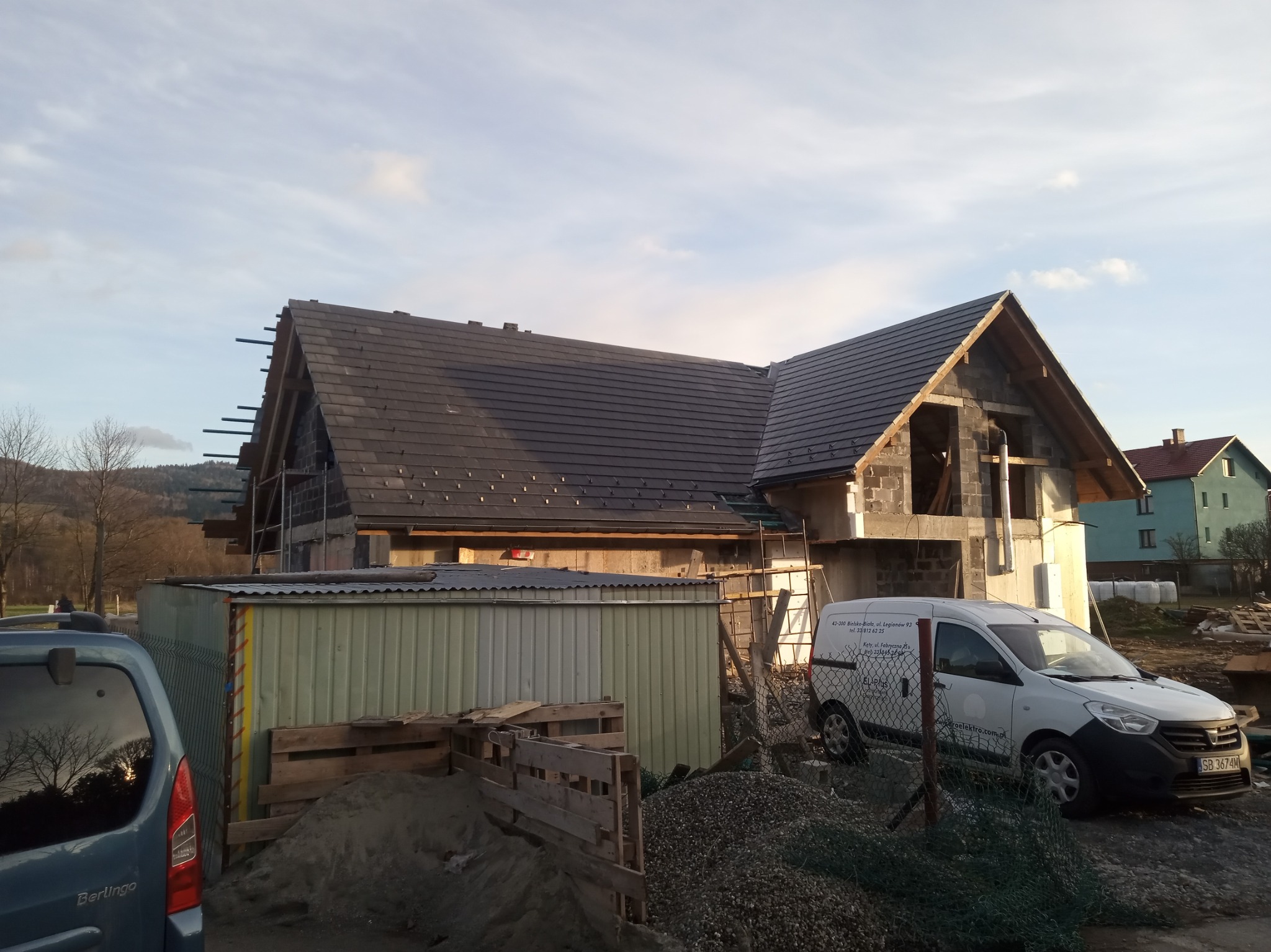 Budowa domu murowanego w trakcie realizacji: widoczna konstrukcja z pustaków, częściowo pokryty dach z ciemnej dachówki, rusztowania, materiały budowlane i samochód dostawczy firmy budowlanej...