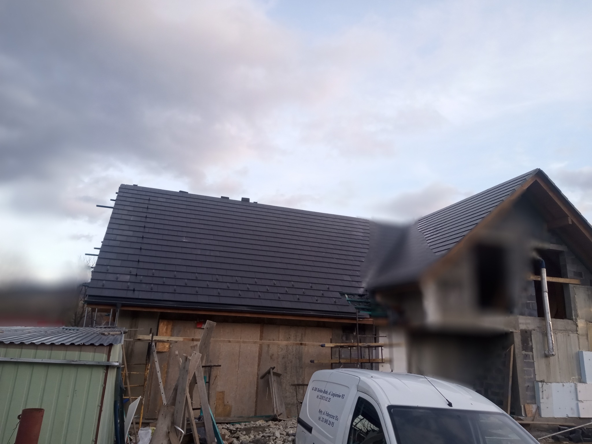 Dom w trakcie budowy z częściowo ukończonym, ciemnym dachem z dachówki ceramicznej, widoczny z boku, na pierwszym planie biały samochód dostawczy z danymi firmy.