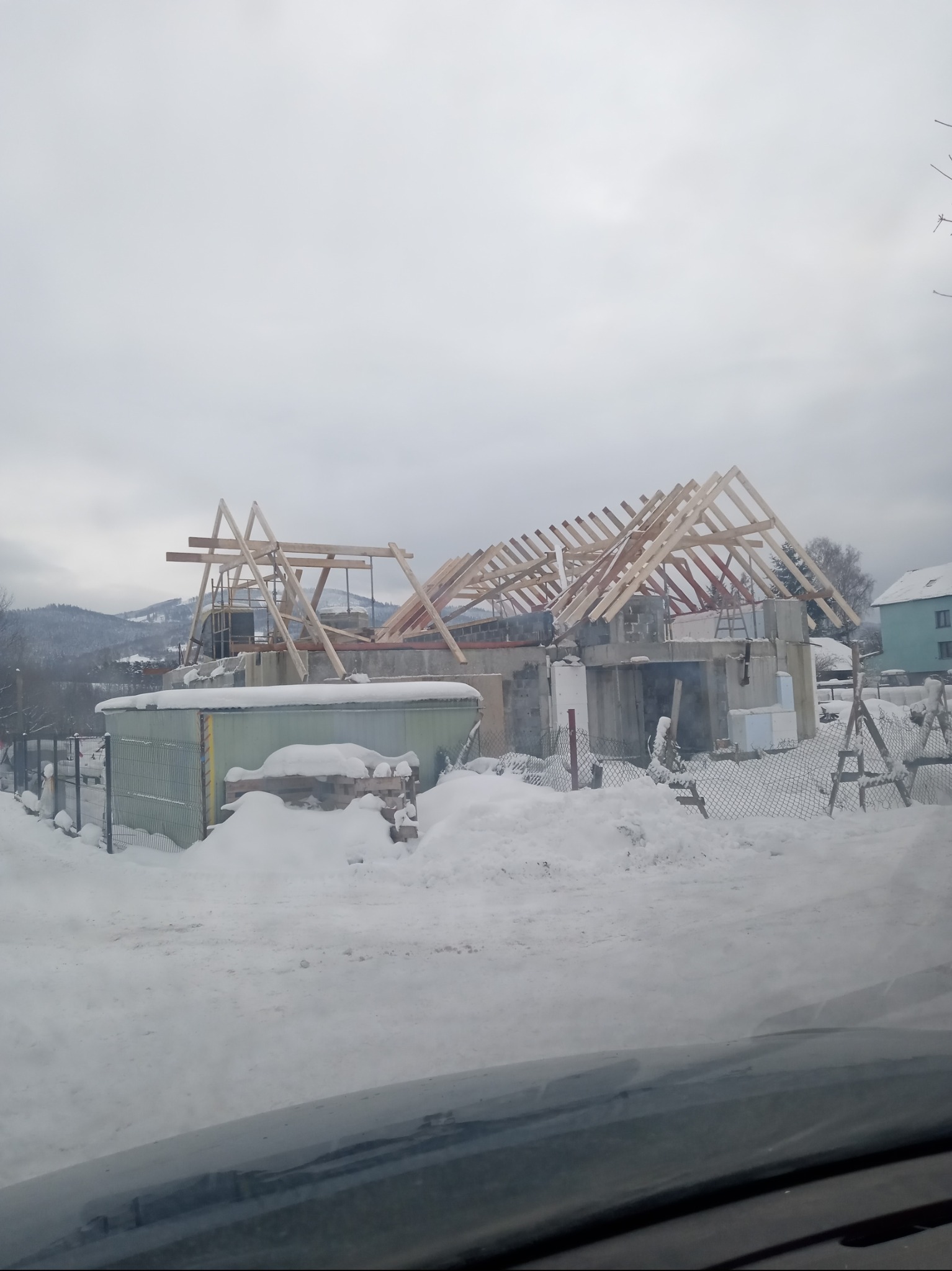 Surowy stan domu murowanego w zimowej scenerii, widoczne drewniane elementy konstrukcji dachu, ośnieżony teren i góry w tle, ujęcie zza szyby samochodu.