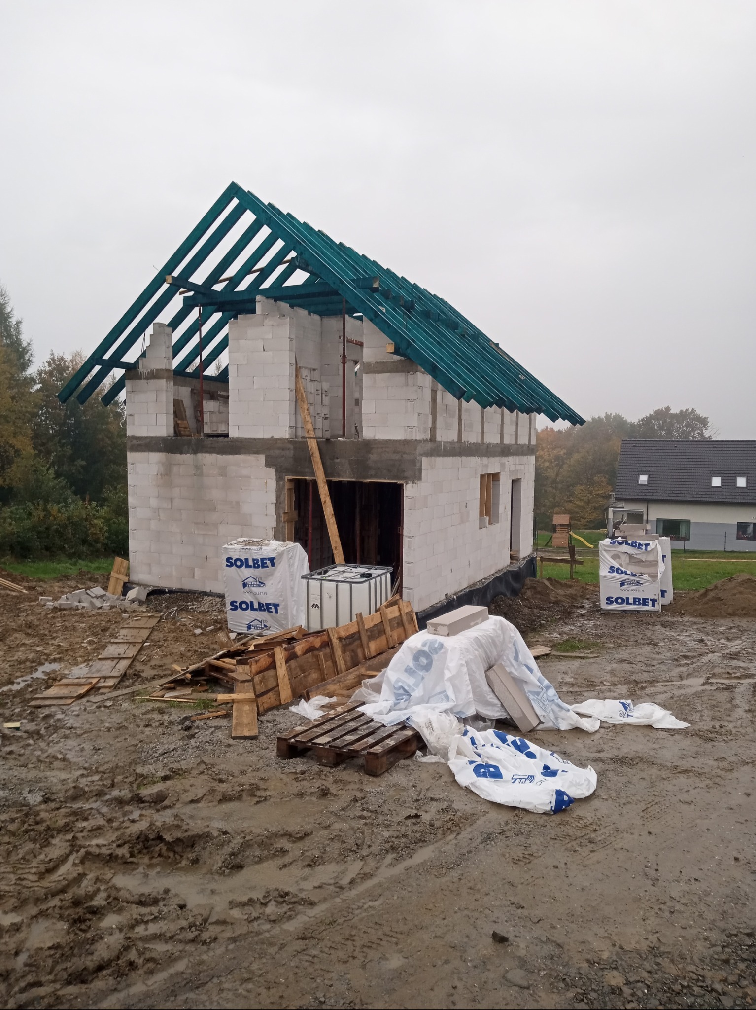 Surowy stan budowy domu z białych bloczków, widoczna zielona konstrukcja dachu, materiały budowlane Solbet i błotniste podłoże.