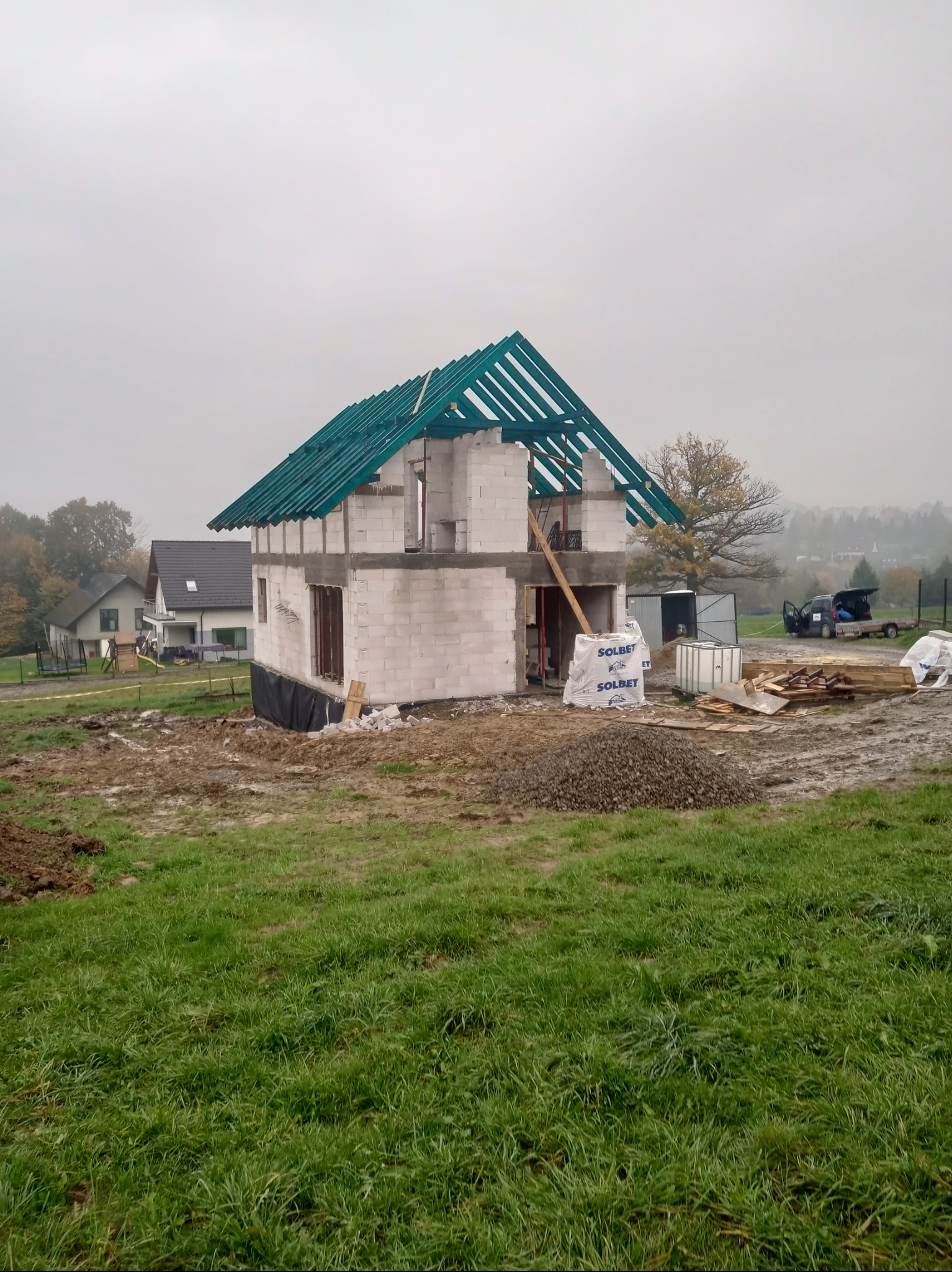 Dom murowany w trakcie budowy, widoczne ściany z bloczków, konstrukcja dachu z zielonymi łatami, materiały budowlane Solbet, teren błotnisty po deszczu.