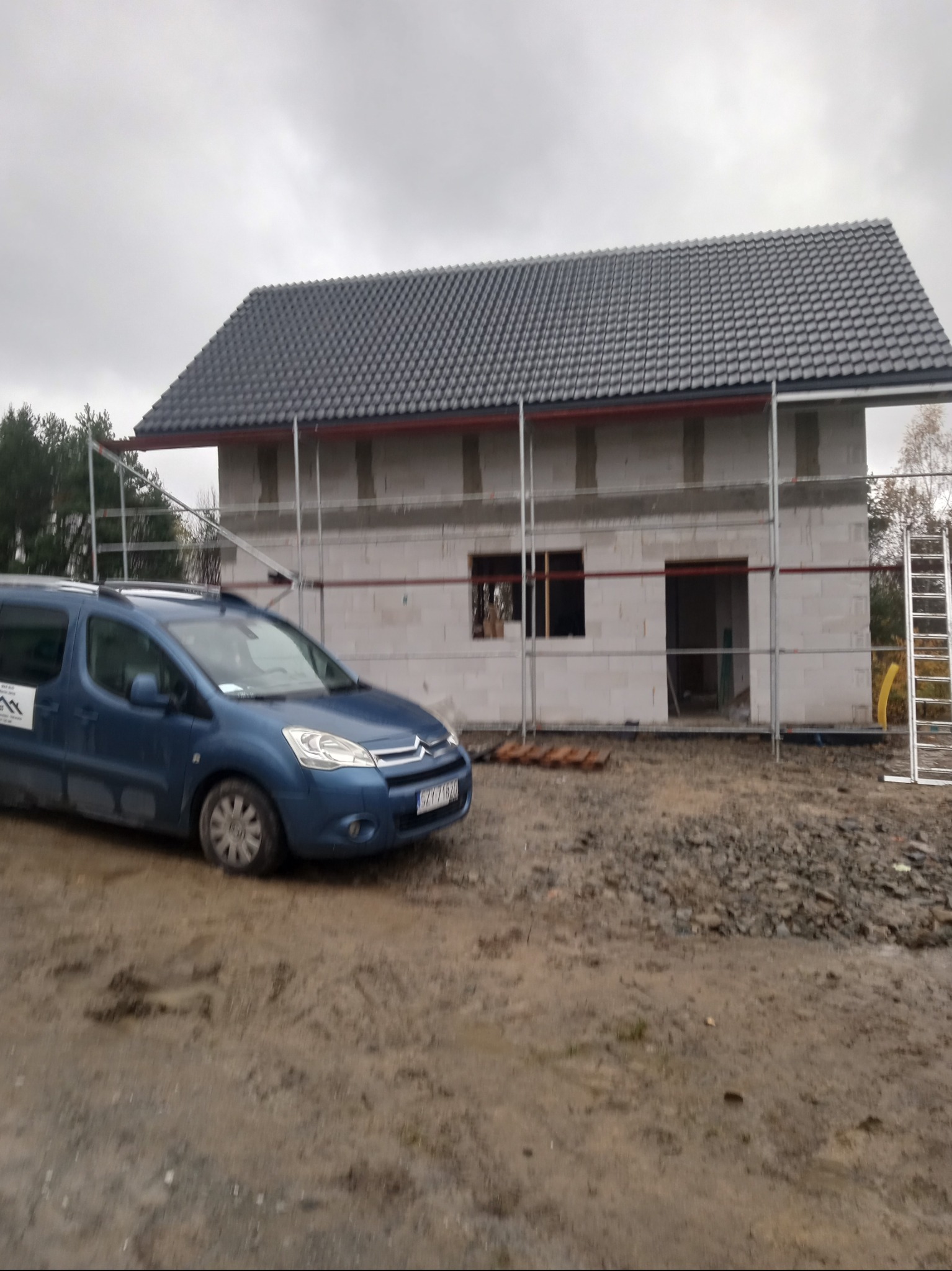 Surowy stan domu murowanego z ciemnoszarą dachówką, otoczonego rusztowaniem, z niebieskim samochodem dostawczym firmy budowlanej zaparkowanym obok na błotnistym terenie.