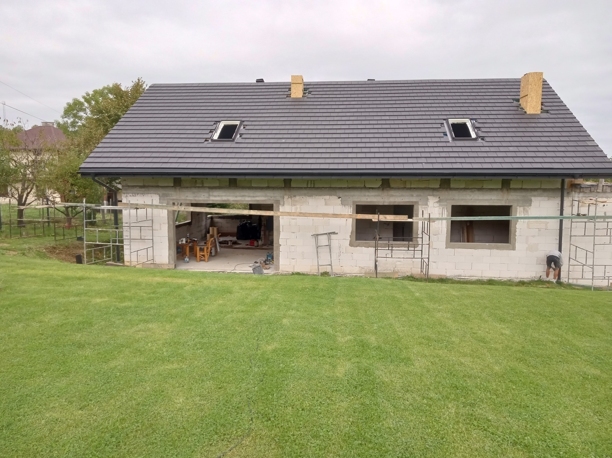 Surowy dom z białego bloczka z nowym, grafitowym dachem z dwoma oknami dachowymi i surowymi kominami, widok z trawnika.