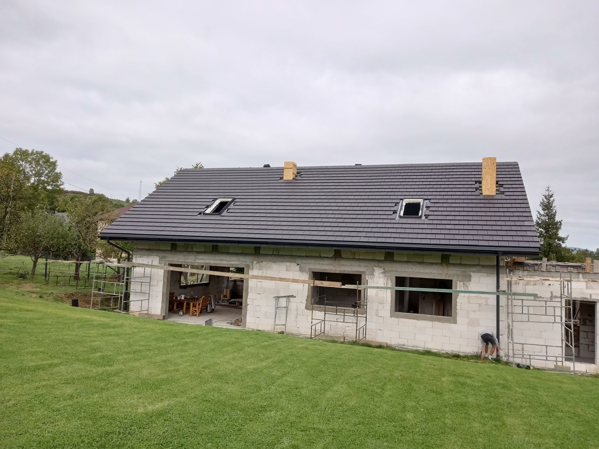 Surowy stan domu z szarego bloczka z ciemnoszarym dachem z dwoma oknami dachowymi i kominem w trakcie budowy, widok z trawnika.