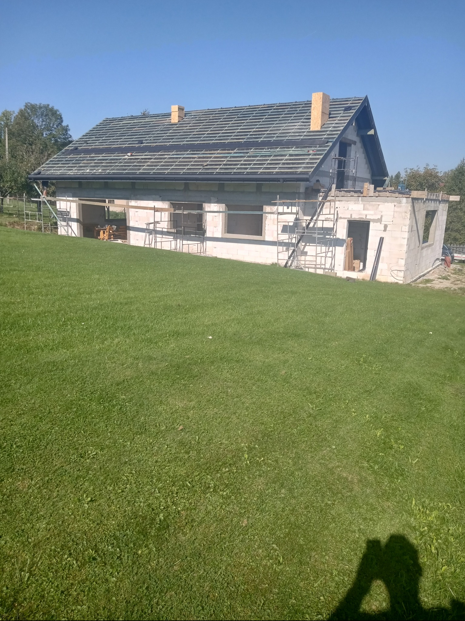 Dom murowany w trakcie budowy z częściowo położonym dachem, widoczne rusztowania i przygotowane krokwie dachowe, na pierwszym planie trawnik.