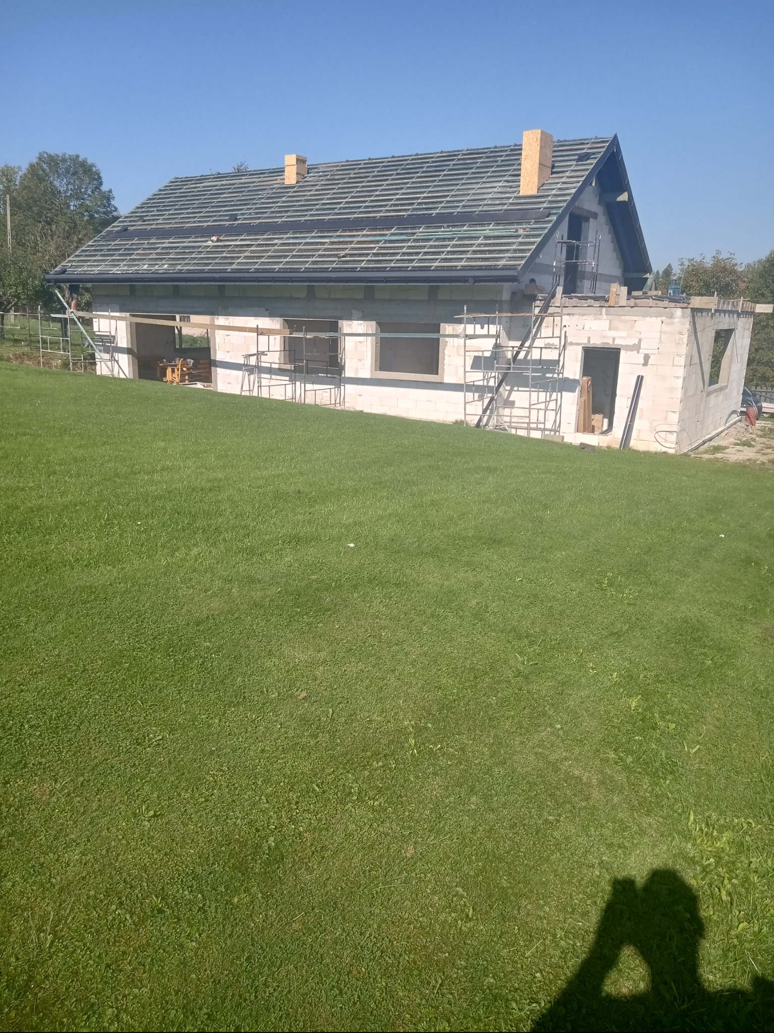 Surowy stan domu murowanego w trakcie budowy, widoczne rusztowania, przygotowana konstrukcja dachu z łatami i kontrłatami oraz kominy w trakcie wznoszenia.