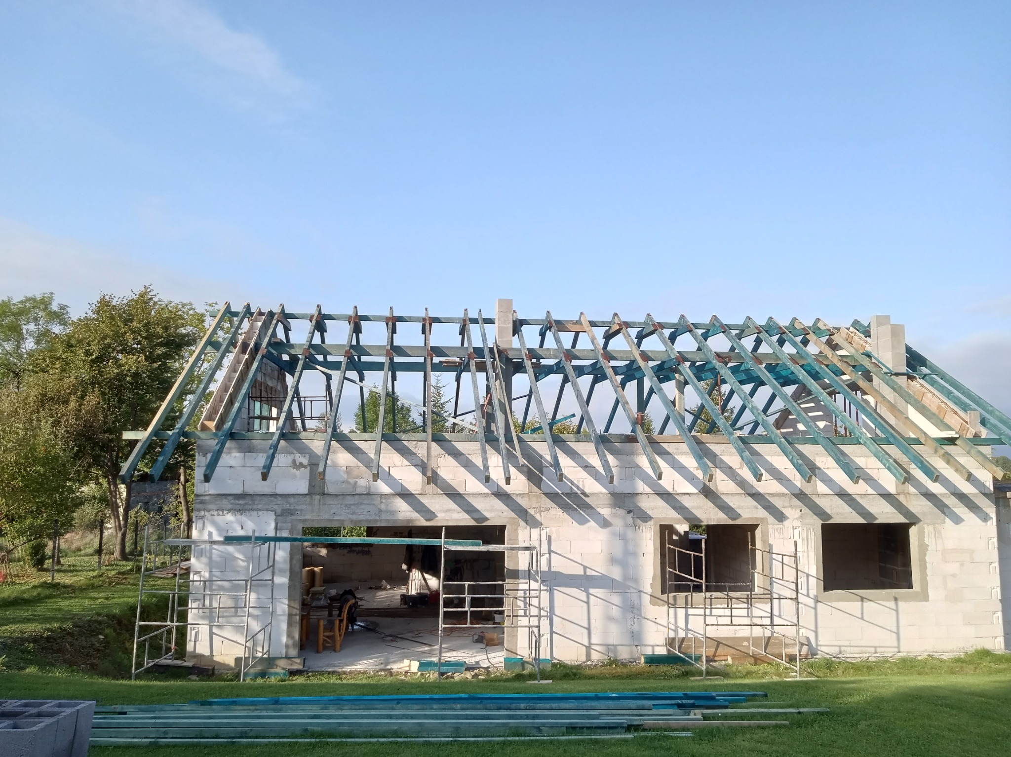 Niedokończony dom z betonu komórkowego w trakcie budowy dachu z drewnianą konstrukcją krokwiową, widoczne rusztowanie i materiały budowlane na trawniku.