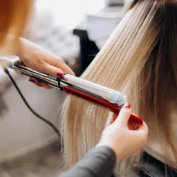 Prostowanie blond włosów przy użyciu prostownicy z czerwonym grzebieniem, widoczne dłonie stylistki z czerwonym manicure.