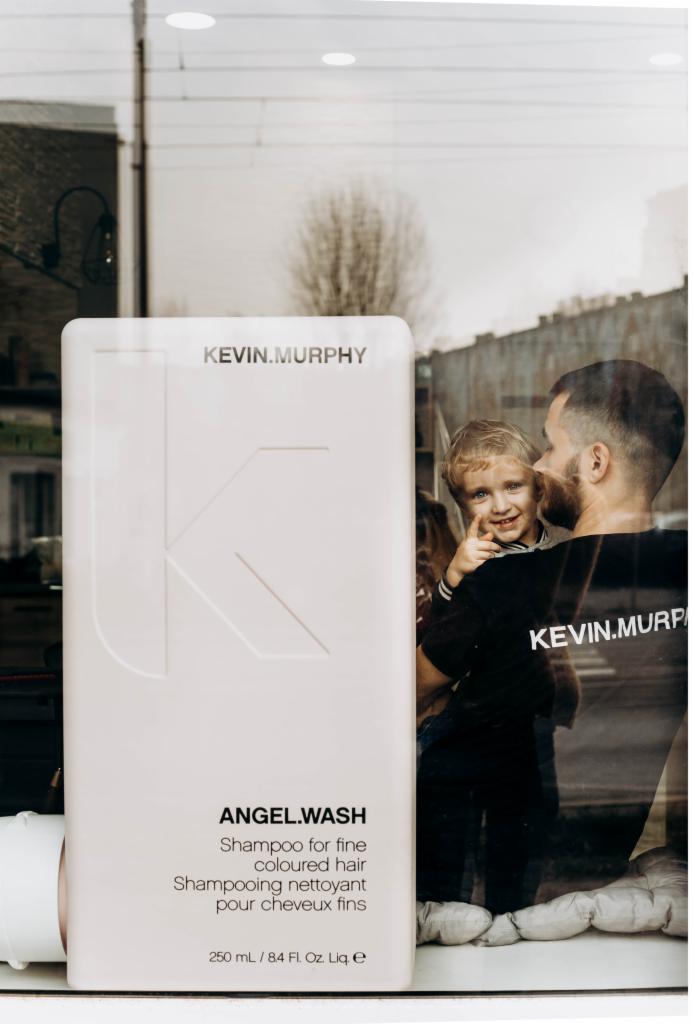 Ekspozycja szamponu Kevin Murphy Angel Wash do włosów farbowanych na tle plakatu z mężczyzną i dzieckiem, widoczna przez szybę.