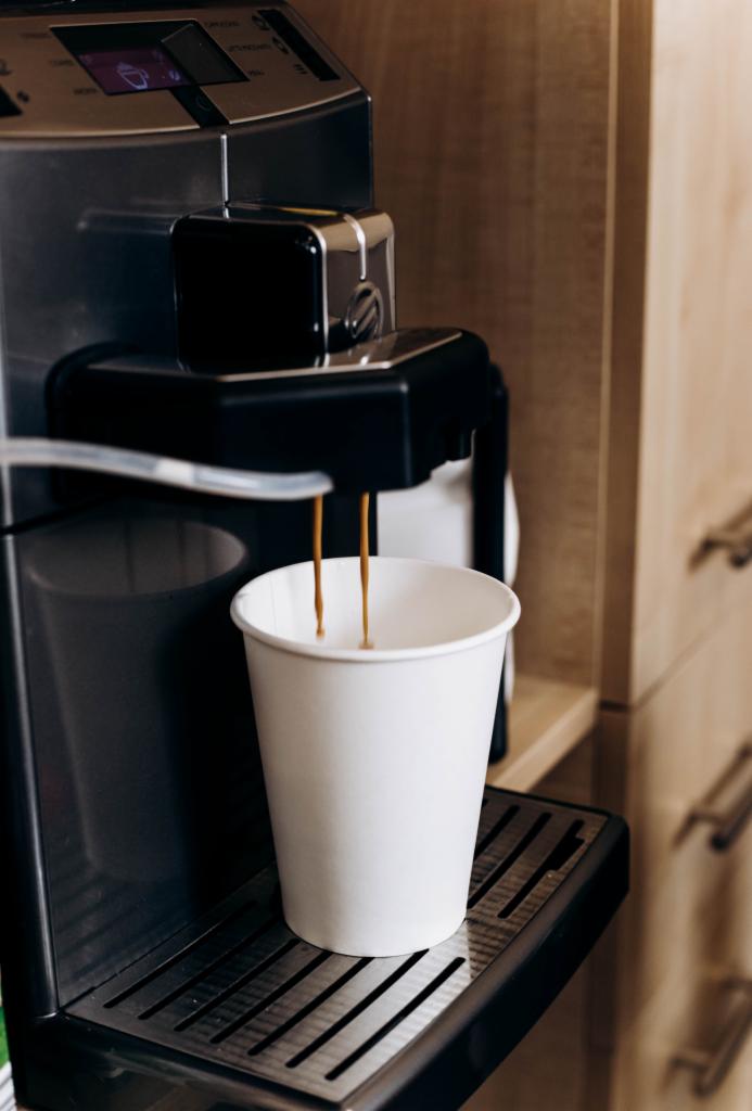 Automatyczny ekspres do kawy nalewa kawę do białego papierowego kubka na tle drewnianych szafek.