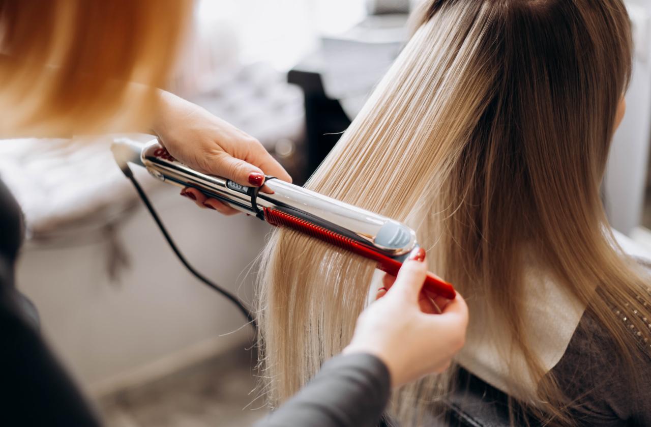 Prostowanie blond włosów przy użyciu prostownicy z czerwonym grzebieniem, widoczne dłonie stylistki z czerwonym manicure.