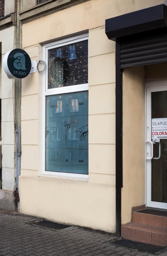 Fasada budynku z oknem ozdobionym grafiką przedstawiającą nożyczki i dłoń, z okrągłym szyldem 'Studio' po lewej stronie i wejściem z napisem 'Olaplex' po prawej.