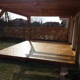 Zadaszony taras z drewnianą podłogą i ażurowym ogrodzeniem, widok na ogród i zabudowania w tle, oświetlony popołudniowym słońcem.