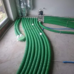 Wnętrze w trakcie montażu instalacji wentylacyjnej z widocznymi zielonymi rurami karbowanymi, srebrnymi przewodami wentylacyjnymi pod sufitem i fragmentem okna.