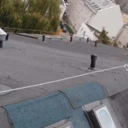 Czarny, płaski dach pokryty papą z widocznymi kominkami wentylacyjnymi, zabezpieczony linką asekuracyjną z karabińczykiem; w tle zabudowa miejska.