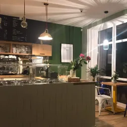 Wnętrze kawiarni w Sade budy z widocznym barem, ekspresami do kawy, menu na ścianie i dekoracjami roślinnymi, oświetlone ciepłym światłem wiszących lamp.