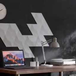 Biuro z ciemną ścianą, na której zamontowane są białe, trójwymiarowe panele dekoracyjne w kształcie strzałek, biurko z laptopem i lampką, zegar na ścianie.