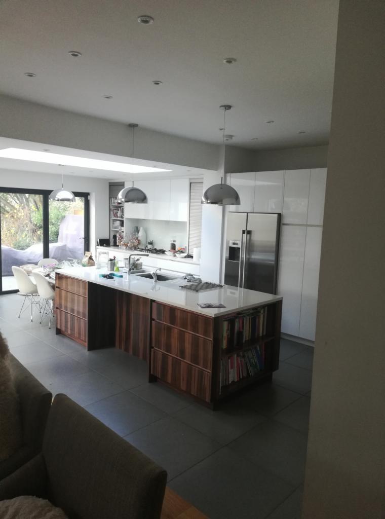 Nowoczesna kuchnia z wyspą o drewnianych frontach, białymi szafkami i stalową lodówką, oświetlona trzema wiszącymi lampami i światłem wpadającym przez okno dachowe.