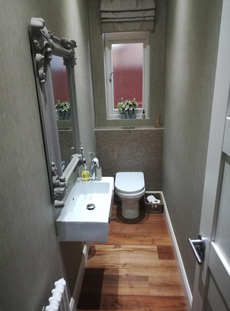 Mała łazienka z drewnianą podłogą, wiszącą umywalką, ozdobnym lustrem i toaletą. Nad toaletą dekoracyjny panel z mozaiki. Na parapecie okna doniczki z kwiatami.