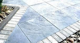 Szare, kwadratowe płyty ułożone na zewnątrz, obramowane jaśniejszą kostką i drobnymi kamieniami, tworzące wzór geometryczny. Na obrzeżach widoczna zieleń roślin. Widok z góry.