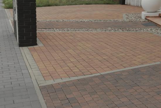 Ułożona kostka brukowa w odcieniach brązu i czerwieni tworząca wzór na podjeździe, oddzielona pasami z jasnego żwiru, z widocznym fragmentem kolumny z ciemnej cegły i szarej kostki po lewej stronie.