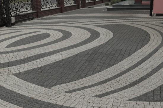 Ułożona nawierzchnia z kostki granitowej w dwóch odcieniach szarości, tworząca wzór koncentrycznych okręgów i prostych linii, prowadząca do schodów i ogrodzenia z dekoracyjnymi elementami.