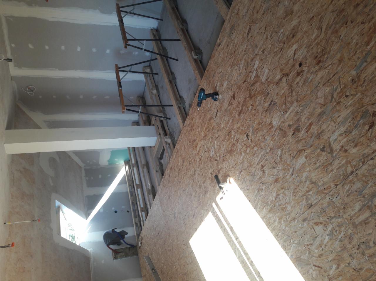 Wnętrze w trakcie prac wykończeniowych: płyta OSB przykręcana wiertarką, widoczne szpachlowane ściany i sufit, fragment okna i pracownik w kasku.