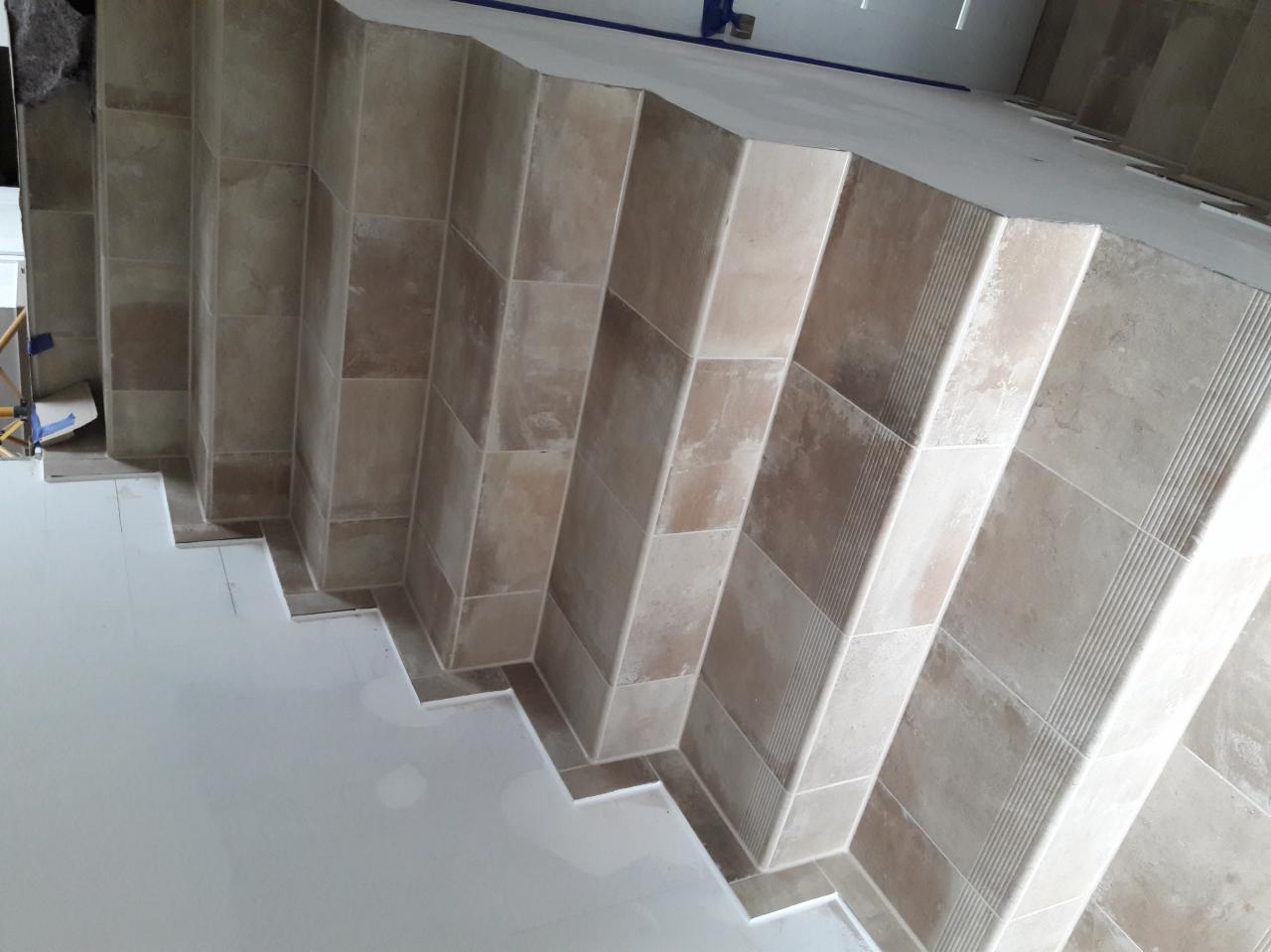 Wnętrze w trakcie wykańczania: schody obłożone płytkami w odcieniach beżu i brązu, z widocznymi detalami wykończeniowymi i fragmentem zabezpieczonej podłogi.
