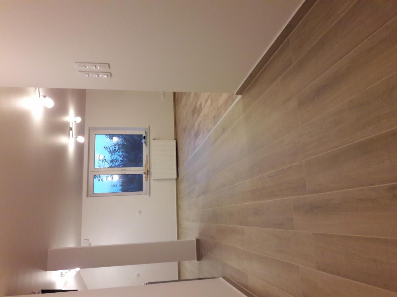 Wykończone wnętrze pokoju z drewnopodobną podłogą, białymi ścianami, grzejnikiem i oknem z widokiem na drzewa.