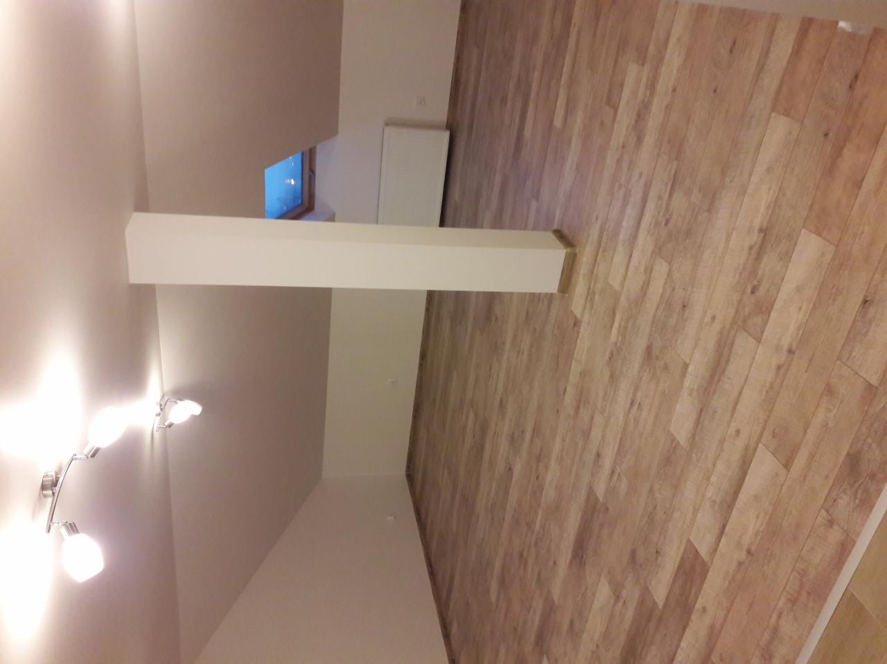 Wykończone wnętrze pokoju z drewnianą podłogą, białymi ścianami, kolumną i lampą sufitową z trzema reflektorami.