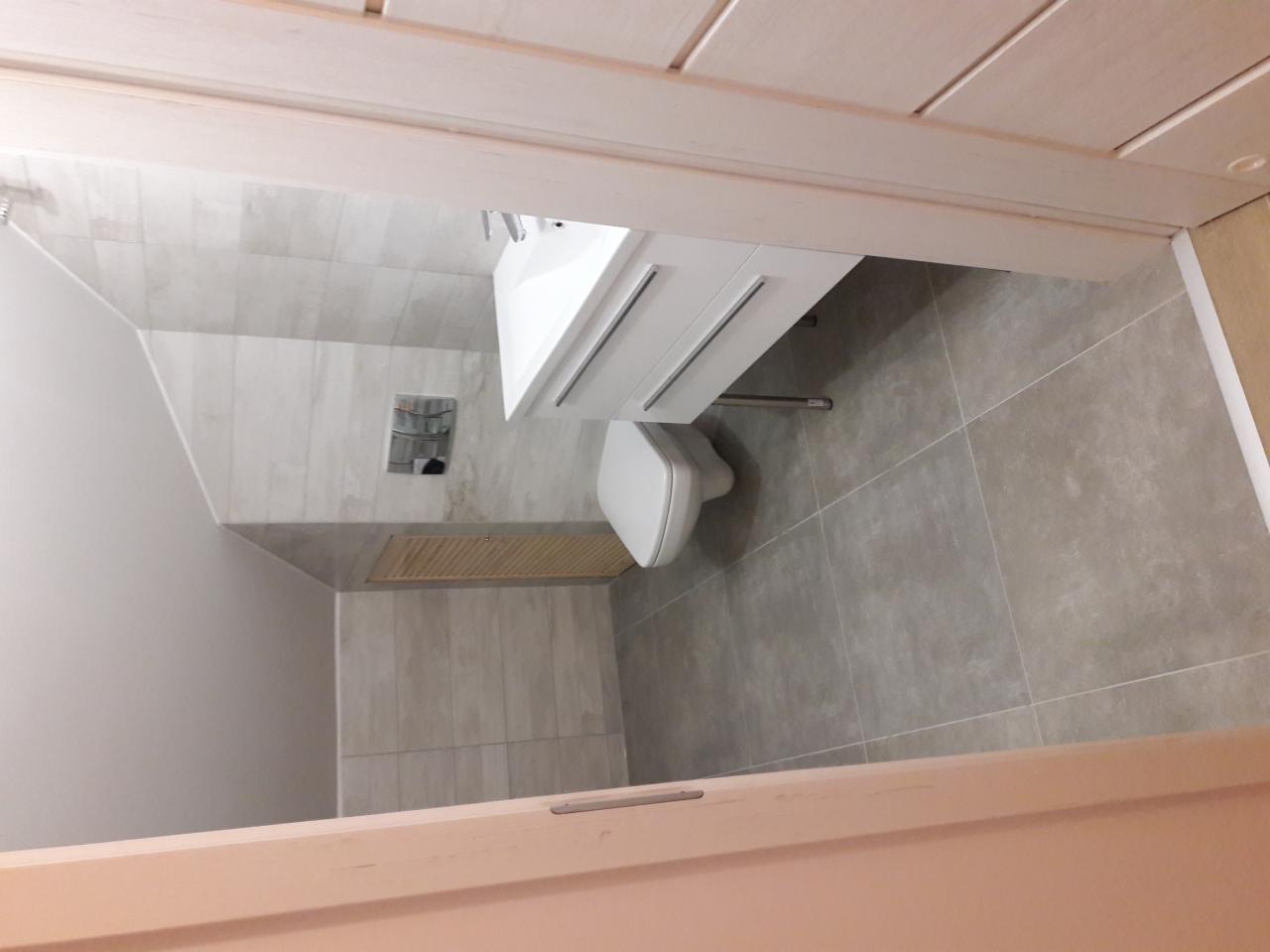 Nowoczesna łazienka z szarymi płytkami, wiszącą toaletą i białą szafką z umywalką, widziana zza częściowo otwartych drzwi.