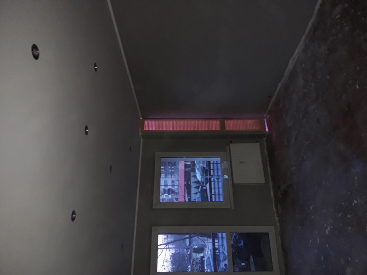 Wnętrze pokoju w trakcie prac wykończeniowych, widoczne szpachlowane ściany, przygotowane do malowania, dwa okna z widokiem na zewnątrz, oraz otwory w suficie pod oświetlenie.