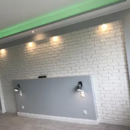 Ściana z białej cegły dekoracyjnej z szarą wstawką i oświetleniem punktowym, wykończona listwami przypodłogowymi, z podświetleniem LED na suficie.