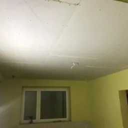 Niedokończony sufit z płyt gipsowo-kartonowych w pokoju z żółtymi ścianami, widoczne łączenia płyt, wystające kable instalacji elektrycznej, okno z widokiem na zewnątrz.