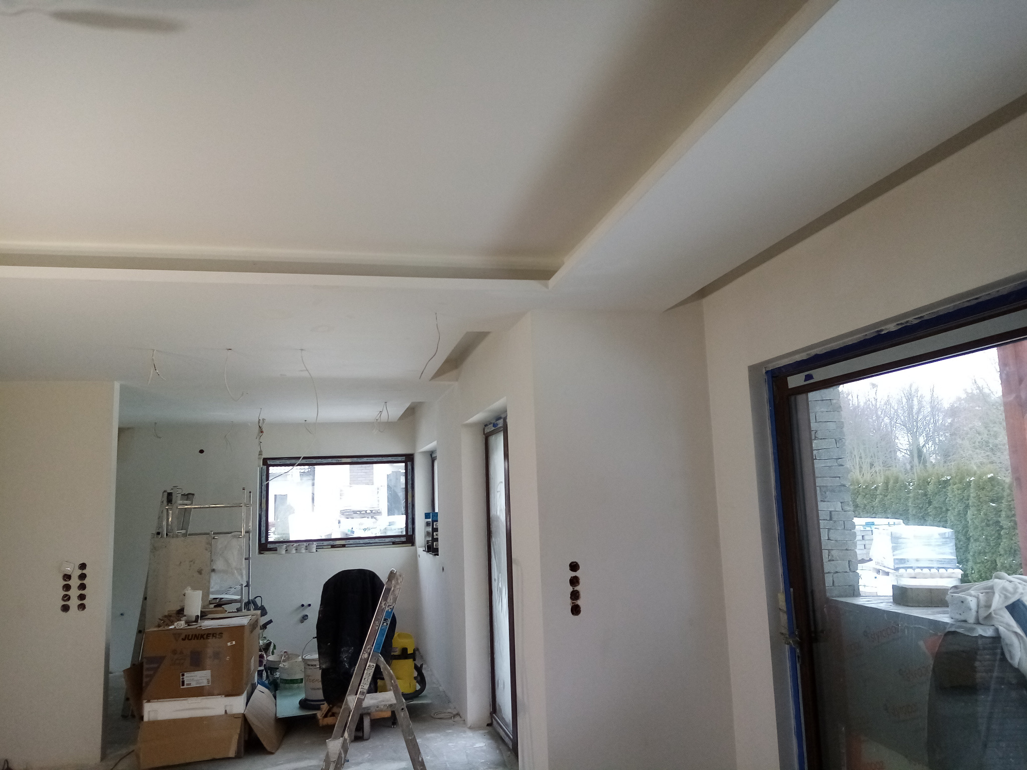 Wnętrze w trakcie prac wykończeniowych: widok na sufit z dekoracyjnymi podcieniami, ściany przygotowane do malowania, okna zabezpieczone folią, widoczny sprzęt budowlany i materiały.