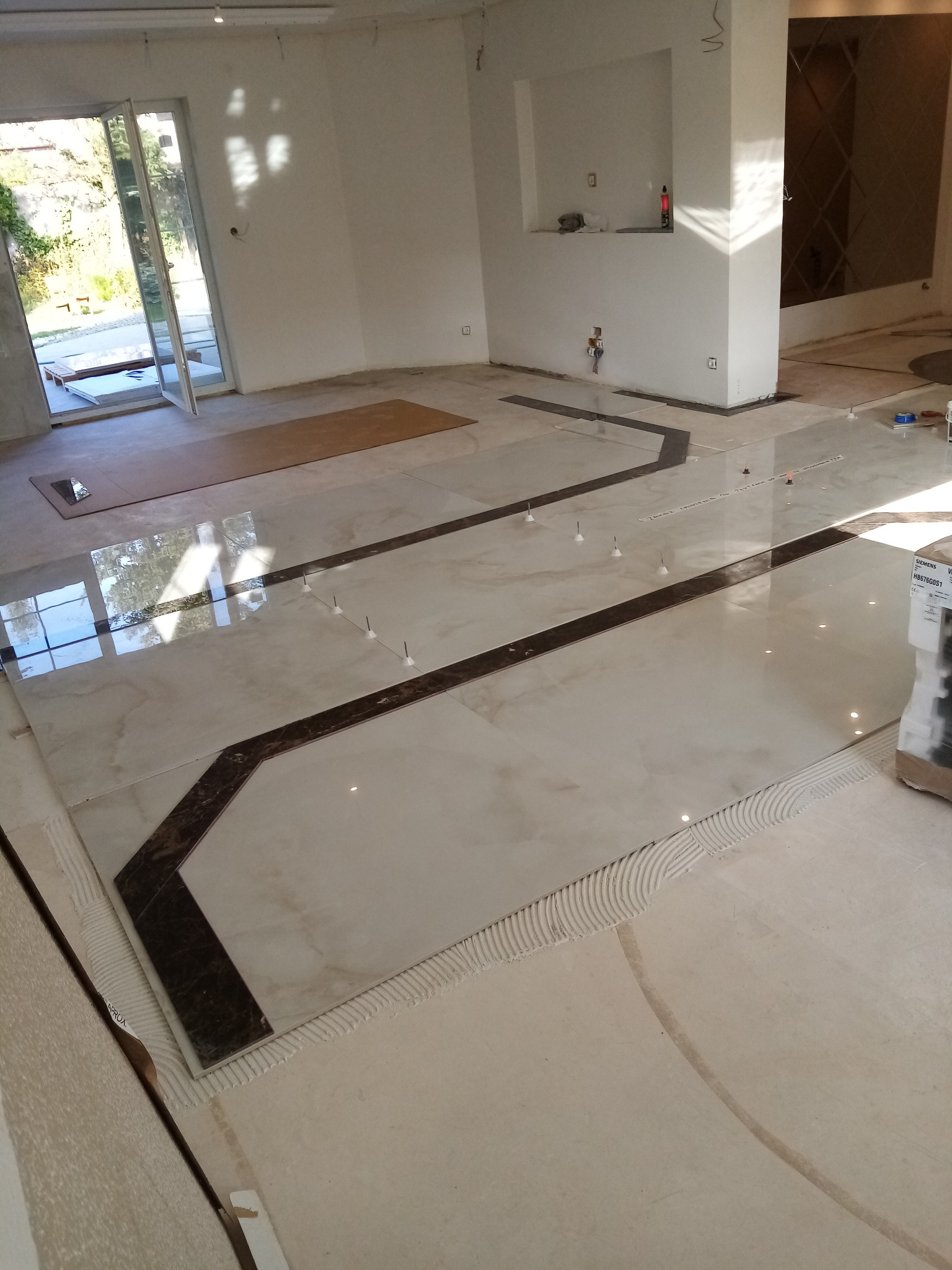 Układanie jasnych, marmurkowych płytek podłogowych z ciemnobrązowymi wstawkami w Poznaniu, widoczne kliny poziomujące i świeży klej, częściowo wykończona podłoga.