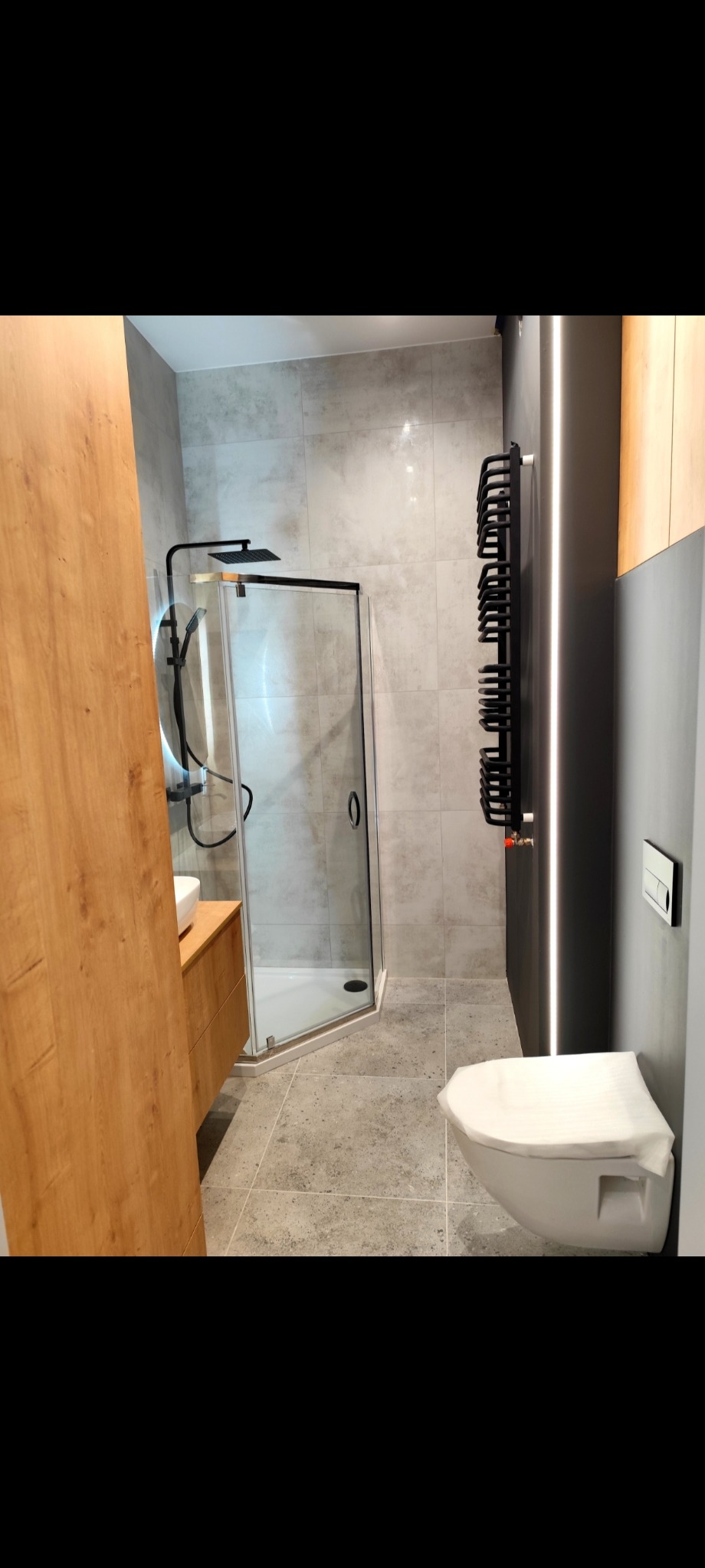 Nowoczesna łazienka z kabiną prysznicową typu walk-in, grzejnikiem drabinkowym w kolorze czarnym i podwieszaną toaletą, wykończona w szarościach z elementami drewna.