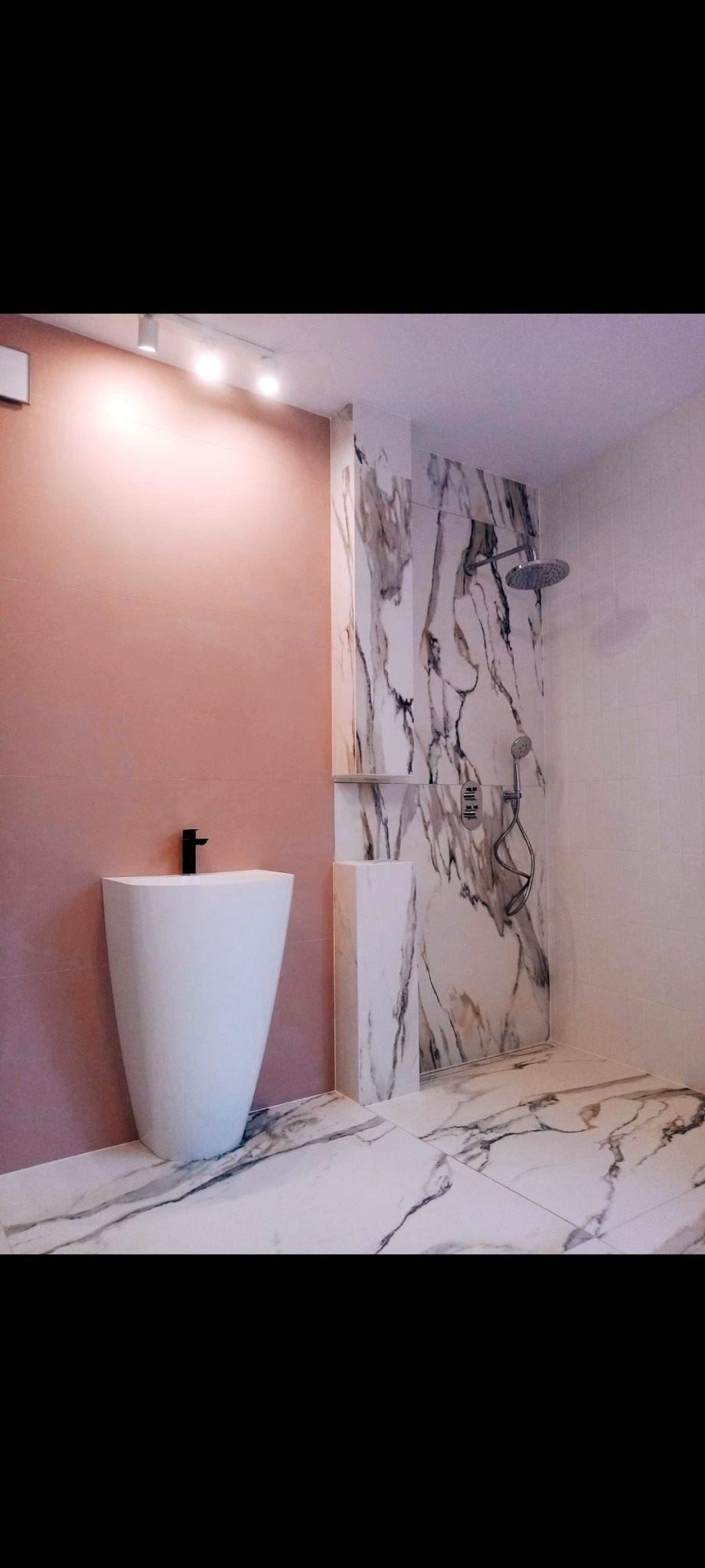 Nowoczesna łazienka z wolnostojącą umywalką, marmurowymi płytkami i różową ścianą, oświetlona punktowymi lampami sufitowymi.