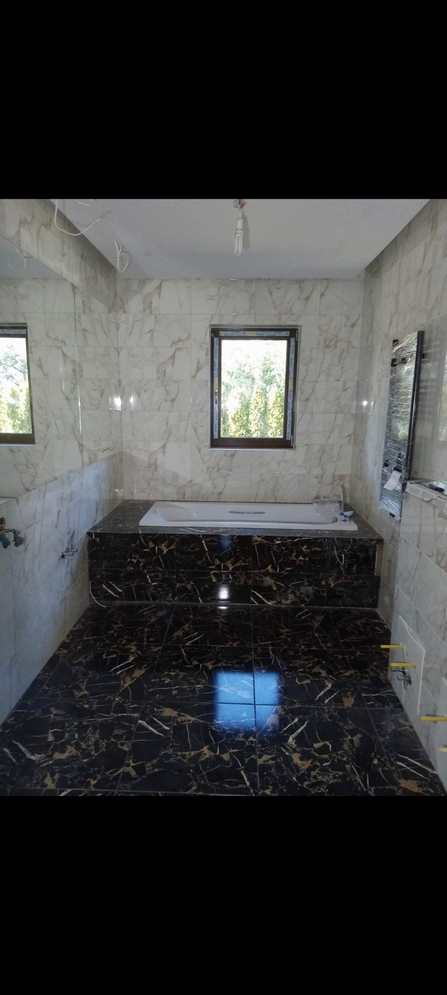 Wykończona łazienka z marmurowymi płytkami na ścianach i podłodze, wanna obudowana czarnym marmurem, okno z widokiem na zieleń, widoczne elementy instalacji hydraulicznej.