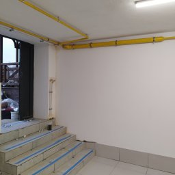 Andrzej Barański - Nowo ułożone żółte rury instalacji gazowej, prowadzone pod sufitem i wzdłuż białej ściany, z widocznymi mocowaniami. Obok niedokończone schody z płytkami i taśmą.