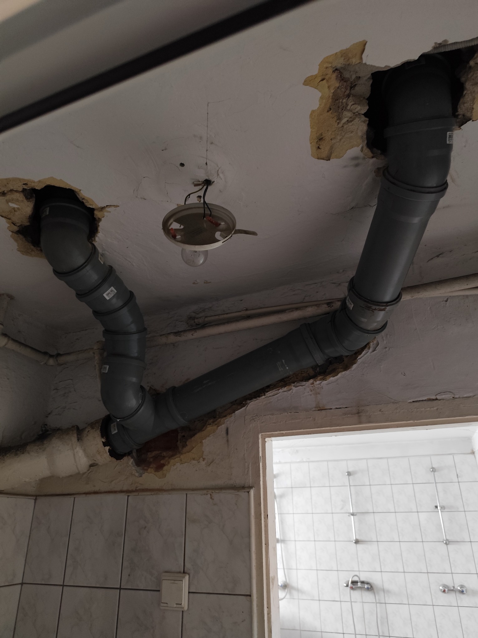 Szare rury kanalizacyjne połączone pod sufitem z widocznymi uszkodzeniami tynku, w tle fragment łazienki z białymi płytkami. Prace hydrauliczne w trakcie wymiany pionu.