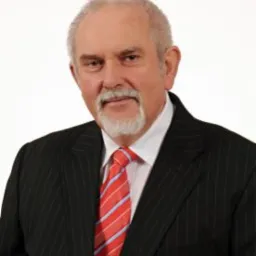 Portret starszego mężczyzny z siwą brodą i wąsami, ubranego w ciemny garnitur w prążki i czerwoną krawat w paski, na jasnym tle.